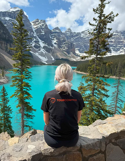 Kvinna med TourCompass T-shirt blickar ut över en turkosblå sjö och snötäckta berg i Kanada