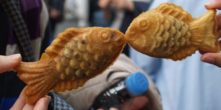 Två personer håller upp taiyaki-kakor så det ser ut som om fiskarna pussas
